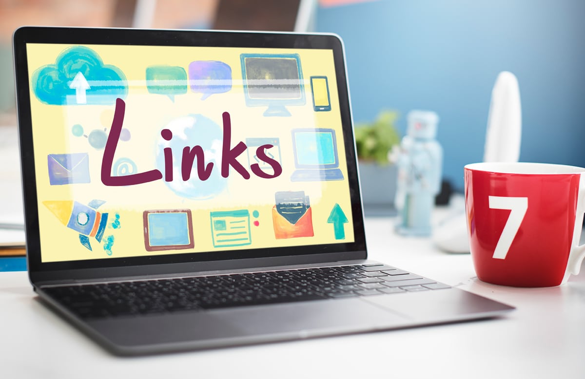 A laptop displaying a colorful screen with the word Links, various icons, and anchor texts, sits next to a red mug labeled 7 on a white desk.