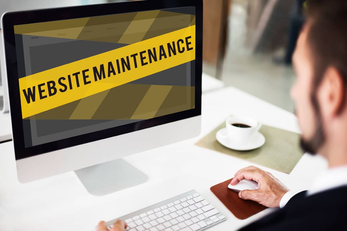 A man in business attire sits at a desk, looking at a computer screen displaying a yellow Website Maintenance banner, using a website checklist to secure your site. A cup and saucer rest on the desk beside him.