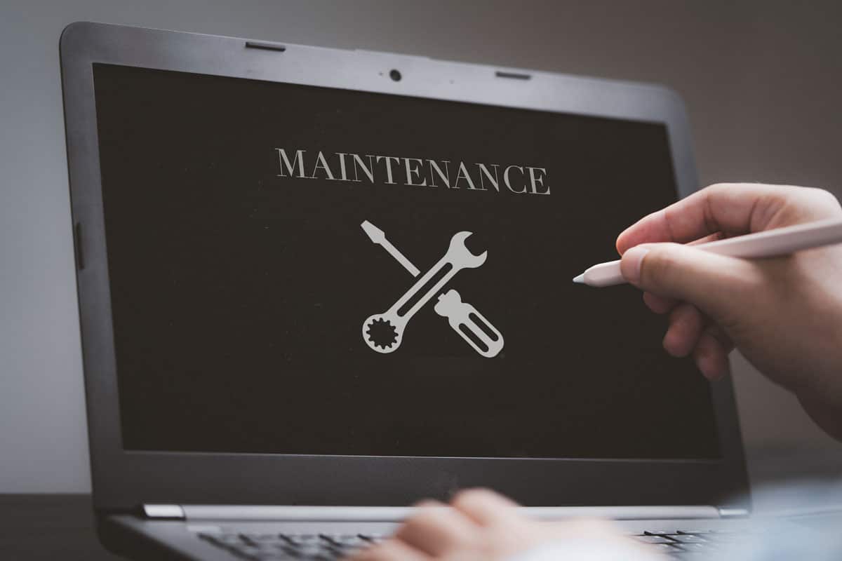 A person uses a stylus on a laptop displaying the word MAINTENANCE above icons of a wrench, screwdriver, and gear, symbolizing repair or technical support—ideal for illustrating small business website maintenance or essential website maintenance tips.